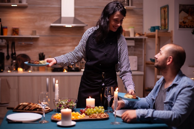 Photo wife surprising husband with dinner in dining room and celebrating with delicious food. woman preparing festive dinner, cooking for his man romantic dinner, talking, sitting at table