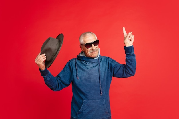 Wijzend roken portret van senior man in stijlvolle brillen en hoed geïsoleerd op rode studio