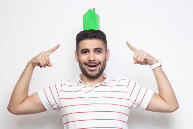 Wil je een eigen huis? Gelukkige jonge man in t-shirt met groenboek klein huis op het hoofd en wijzende vinger naar het plannen van een nieuw huis om te kopen. Binnen, geïsoleerd, studio-opname, kopieer ruimte, witte achtergrond