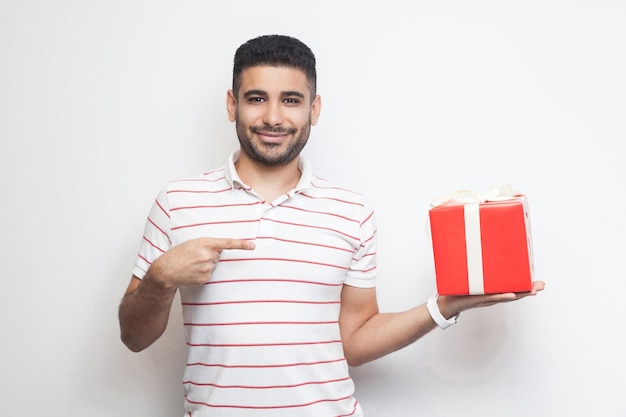 Wil jij? Portret van een vrolijke jonge volwassen man in een t-shirt met een rode geschenkdoos en een wijzende vinger, kijkend naar de camera met een glimlach. Binnen, geïsoleerd, studio-opname, kopieer ruimte, witte achtergrond