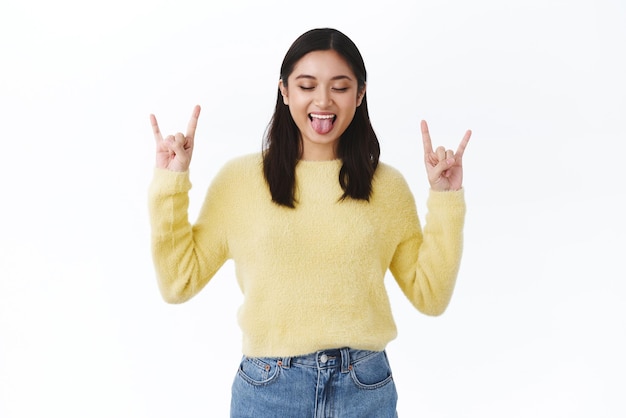 Wild and free millennials Gorgeous carefree asian girl in yellow sweater having fun at awesome student party close eyes show tongue and smiling make rock n roll gesture to express excitement