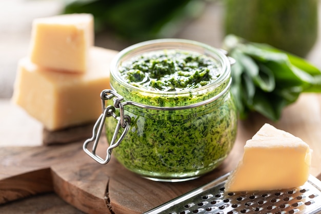 Wild leek pesto with olive oil and parmesan cheese in a glass jar on a wooden table. Useful properties of ramson. Leaves of fresh ramson. Horizontal