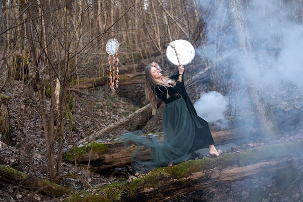 Foto donna selvaggia che suona il tamburo sciamanico in piedi sul tronco di un albero di muschio verde nella foresta bomba di fumo bianco