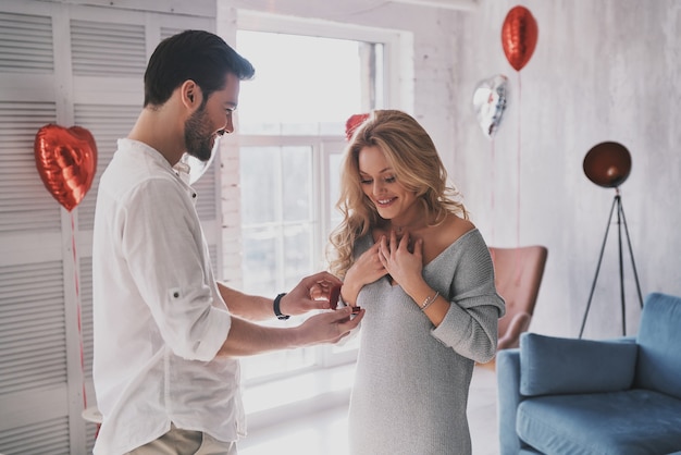 Will you marry me? Surprised young woman looking at engagement ring with smile while standing in the bedroom full of balloons