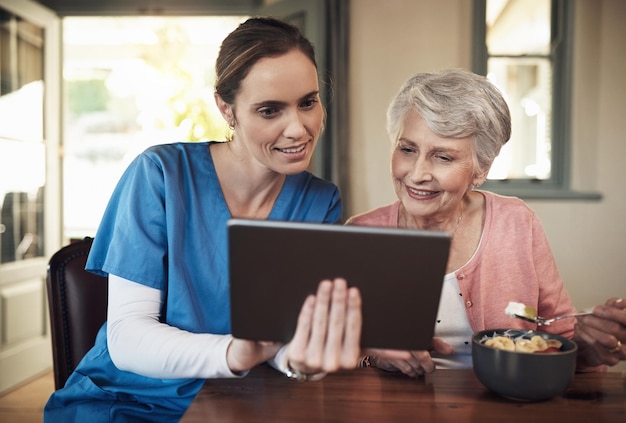 Wilt u uw familie een foto sturen van een jonge verpleegster en oudere vrouw die een digitale tablet gebruiken tijdens het ontbijt in een verpleeghuis