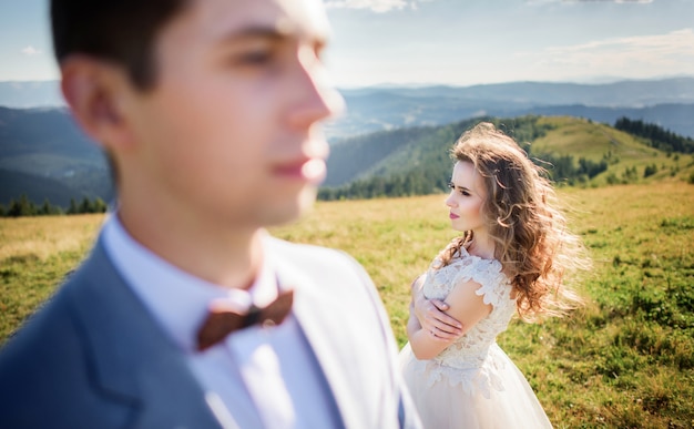 Photo wind blows bride's hair while she stands behind groom on the hill