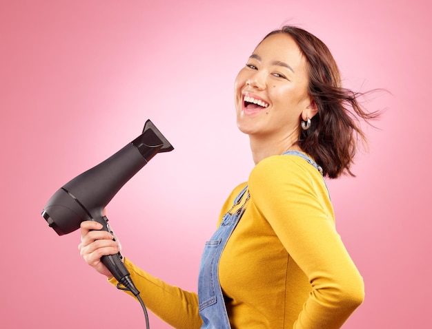 Wind salon and hair care with woman and hairdryer in studio for hairdresser beauty and cosmetics Shampoo smile and hairstyle with portrait of asian person on pink background for product and spa