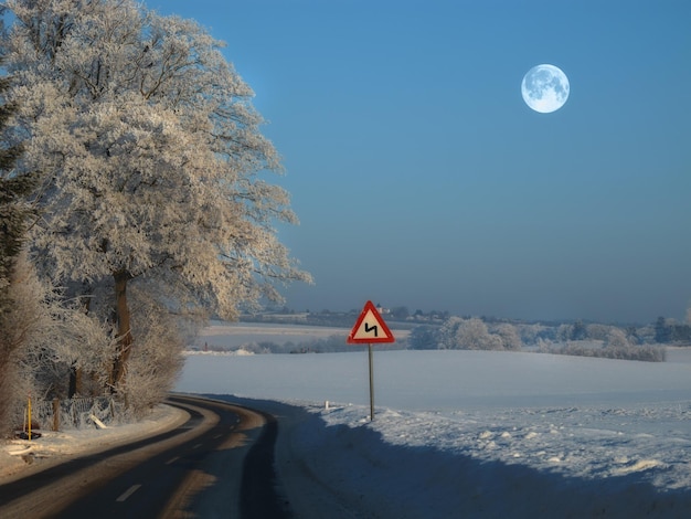 The winding road on a rural winter landscape with a sign on the side of the empty road for safety A slippery and wet road surrounded by snow on a cold winter evening near a forest with ice frost