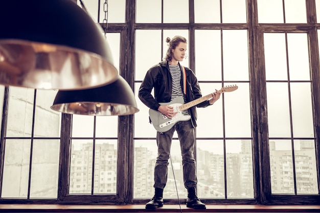 Window sill. Rock musician wearing leather jacket standing on window sill and playing the guitar