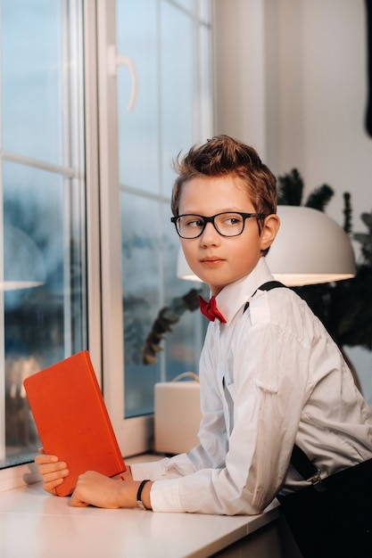 窓際には、赤い蝶ネクタイと眼鏡をかけたシャツを着た男の子が、赤い本を手に持って立っています。