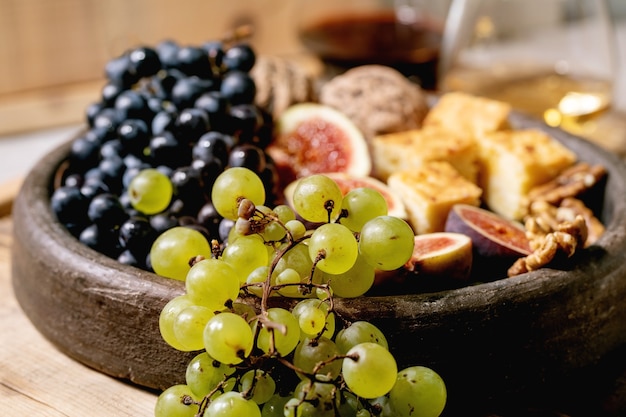 Wine appetizers with different grapes, figs, walnuts, bread, honey and goat cheese on ceramic plate, serving with glasses of red and white wine over old wooden background. close up