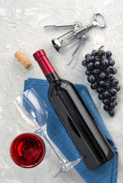灰色のテクスチャ背景にワインボトル、ワイングラス、ブドウ、金属製の栓抜き。上面図。