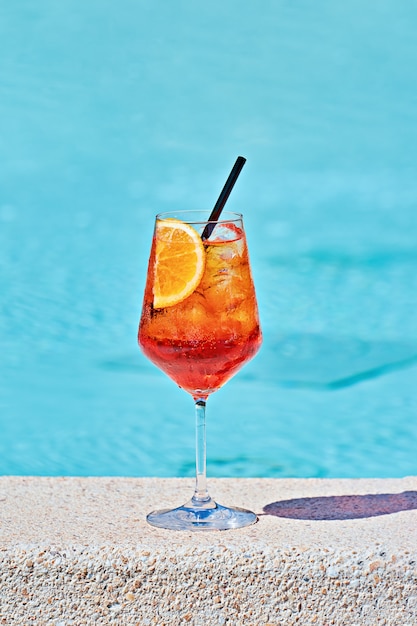 Wine glass of cold cocktail aperol spritz against turquoise\
water of poolside