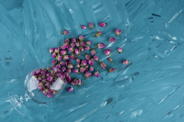 Wineglass full of dried rose flower buds lying on blue table .