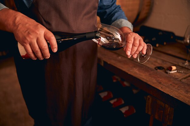 Photo winemaker in apron pouring alcoholic drink into wineglass