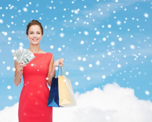 Winkel-, verkoop-, kerst- en vakantieconcept - glimlachende elegante vrouw in rode jurk met boodschappentassen en dollars over blauwe lucht met wolken en sneeuwachtergrond