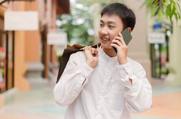 Winkelconcept De winkelende man die boodschappentassen op zijn rug zet terwijl hij met een glimlach aan de winkel aan de rechterkant een gesprek voert met iemand aan de telefoon.