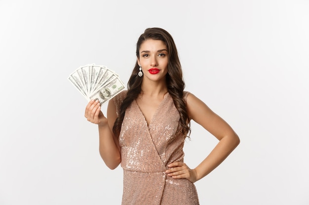 Winkelconcept. Sassy mooie vrouw met rode lippenstift, glamour jurk dragen, geld dollars tonen en zelfverzekerd kijken, staande op een witte achtergrond.