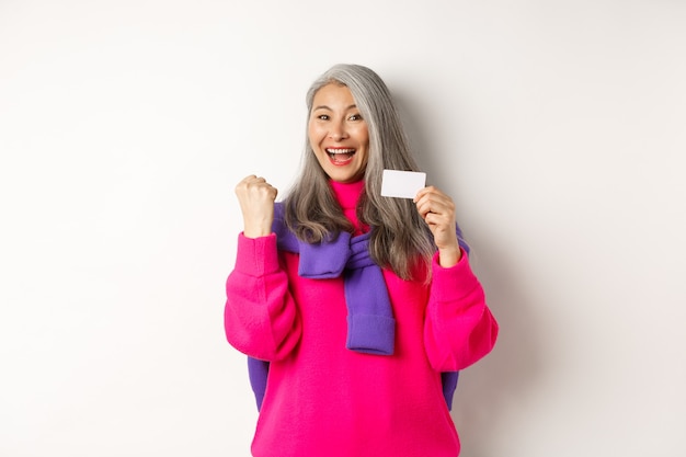 Winkelconcept. Vrolijke aziatische senior vrouw die prijs wint van de bank, met een vuistpompgebaar en plastic creditcard, die gelukkig staat op een witte achtergrond.