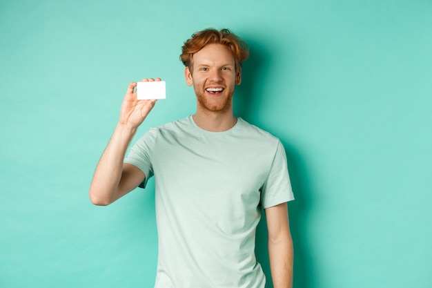 Winkelconcept. Vrolijke jonge man in t-shirt met plastic creditcard en glimlachend, staande over mint achtergrond