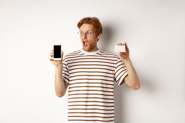 Winkelen en Financiën concept. Verbaasde jonge man met rood haar met een plastic creditcard en een leeg scherm van een smartphone, starend naar het display onder de indruk, witte achtergrond