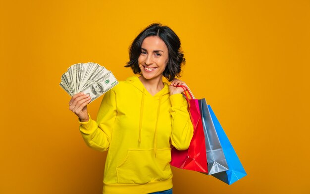 Winkelen was geweldig! succesvol meisje in een gele hoodie, die breed lacht op een camera, haar geld in de ene hand laat zien en aankopen in de andere.