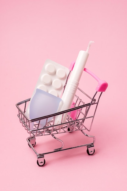 Winkelwagen met menstruatieperiode accessoires op roze achtergrond maandverband Tampons menstruatiecup en pillen verticaal