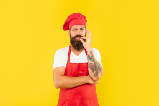 Winking man in red apron and toque licking finger yellow background chef