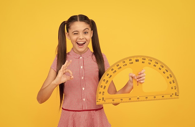 Winking teen girl hold school math tool protractor ruler on yellow background show ok education