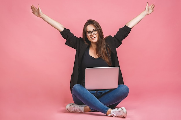 Winnaar bedrijfsconcept. Het portret van gelukkige vrouw in toevallige zitting op vloer in lotusbloem stelt en laptop houdt die over roze achtergrond wordt geïsoleerd