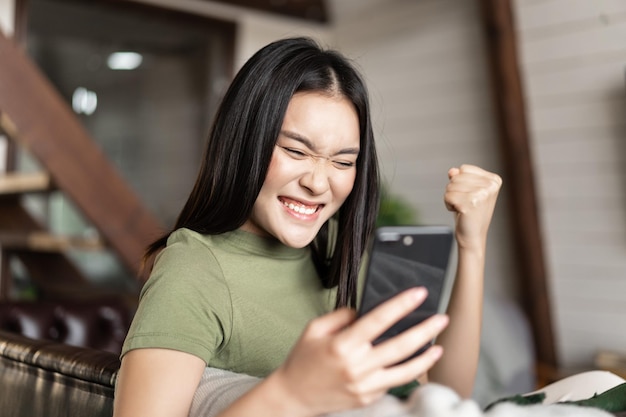 Winnend aziatisch meisje dat thuis zit en naar de mobiele telefoon kijkt, juicht en viert van goede ne...