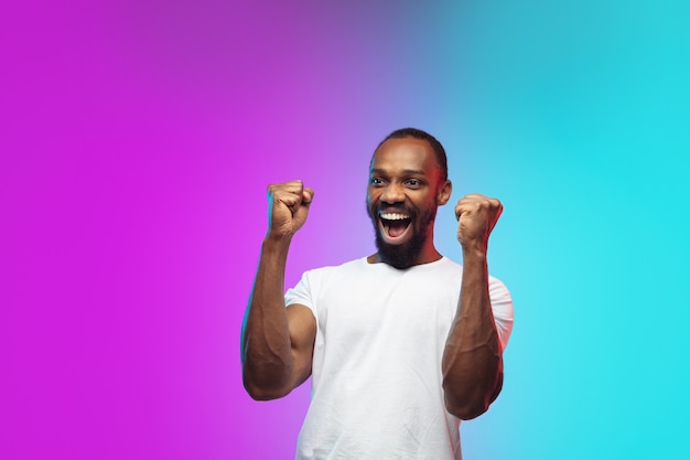 Winner. African-american young man's portrait on gradient studio background in neon. Beautiful male model in casual style, white shirt. Concept of human emotions, facial expression, sales, ad.
