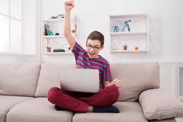 Winner. Happy teenage boy excited at seeing good exam results online. Youngster in cozy home interior