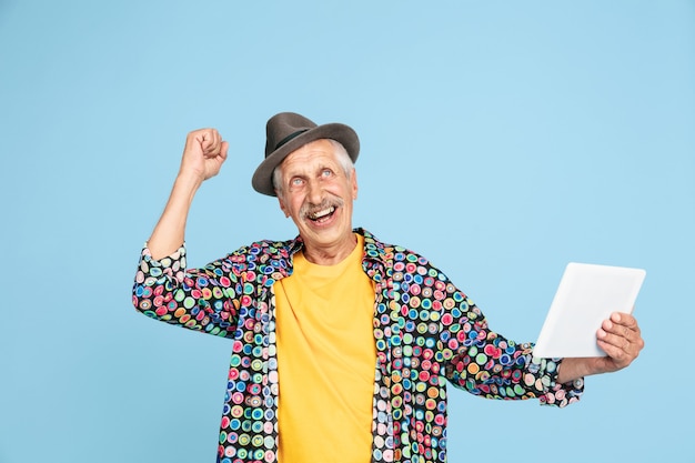 Foto vincitore che tiene tablet ritratto di un uomo anziano hipster con un cappello elegante isolato su studio blu