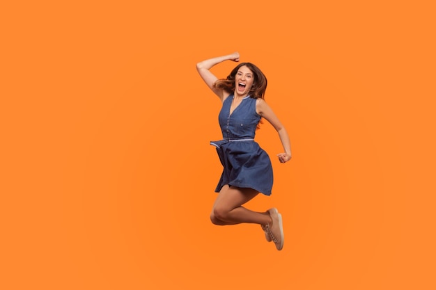 Winner joy Full length of energetic vivid brunette woman in denim dress flying in air with inspired ecstatic expression celebrating great success indoor studio shot isolated on orange background