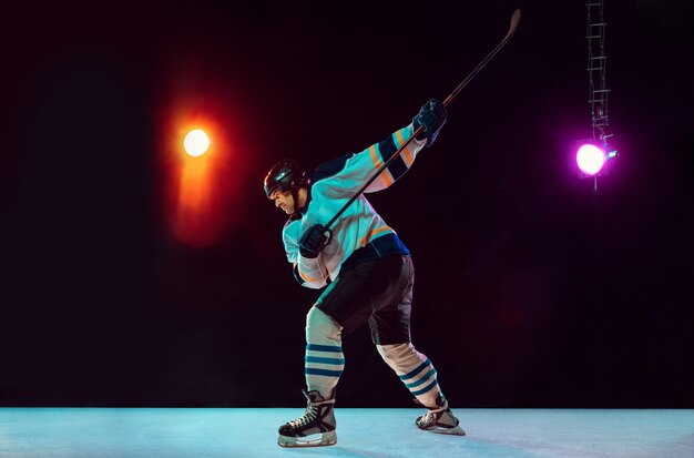 Winner. Male hockey player with the stick on ice court and dark neon colored background. Sportsman wearing equipment, helmet practicing. Concept of sport, healthy lifestyle, motion, wellness, action.