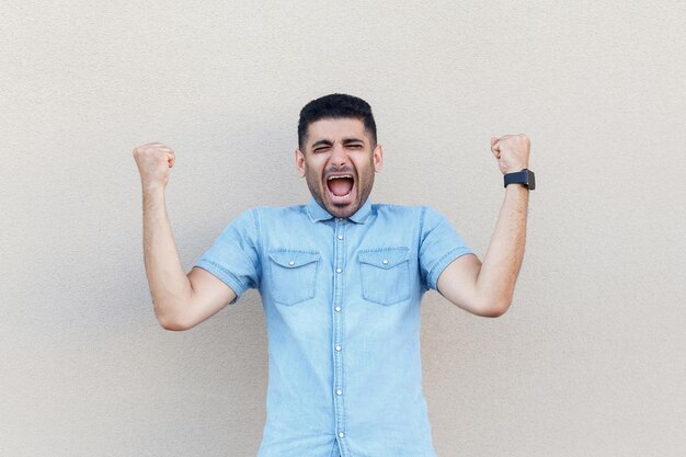 Winner man rejoicing. Portrait of surprised handsome young bearded businessman in blue shirt standing, screaming and celebraiting victory. indoor studio shot isolated on light beige wall background.