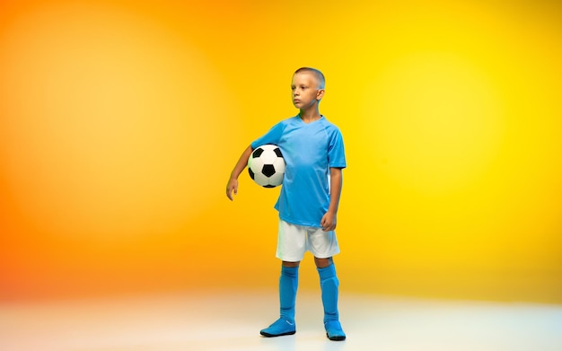 Победитель. молодой мальчик, как футболист или футболист в спортивной одежде, практикующий на градиентно-желтом студийном фоне в неоновом свете. подходящий играющий мальчик в действии, движении, движении в игре. копипространство.