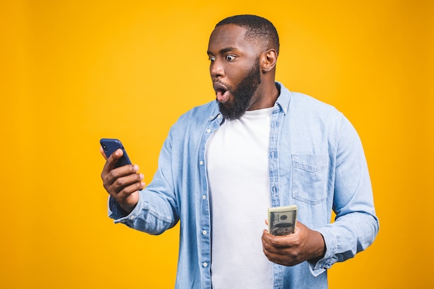 Winner! Young rich african american man in casual t-shirt holding money  dollar bills and mobile phone with surprise