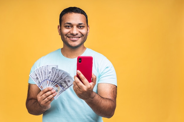 Winner! Young rich happy african american indian black man in casual holding money dollar bills with surprise isolated over yellow wall background. Using mobile phone.