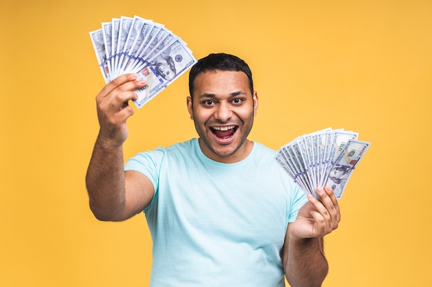 Winner! Young rich happy african american indian black man in casual holding money dollar bills with surprise isolated over yellow wall background.