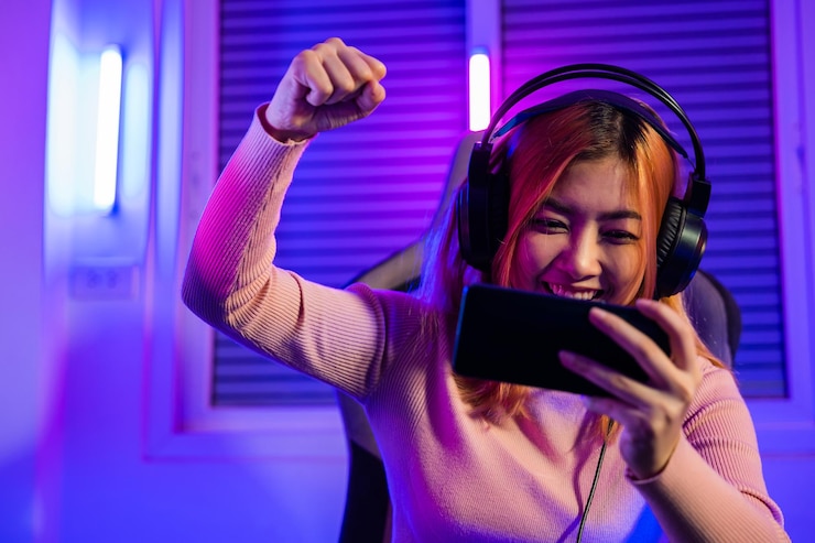 Photo winning happy gamer playing video game online with smartphone with neon lights she raises hands to wins celebrating confused asian woman wearing gaming headphones feeling excited at home