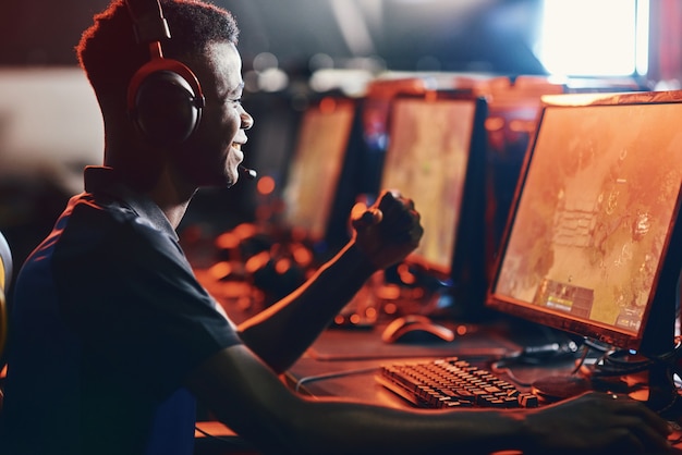 Vincente. vista laterale del ragazzo africano eccitato, giocatore professionista maschio che indossa le cuffie che celebra il successo durante la partecipazione al torneo esport. persone che giocano a videogiochi online