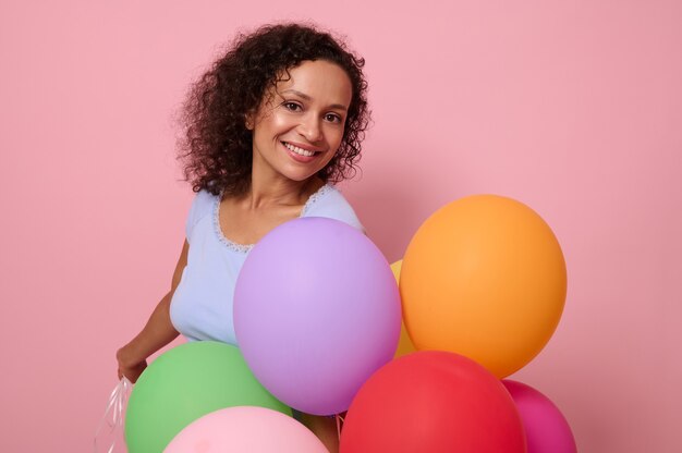 Winsome beautiful mixed race woman with a charming toothy smile, has happy reaction holding bright colorful helium balloons , celebrates birthday, looks at camera, isolated on pink colored background