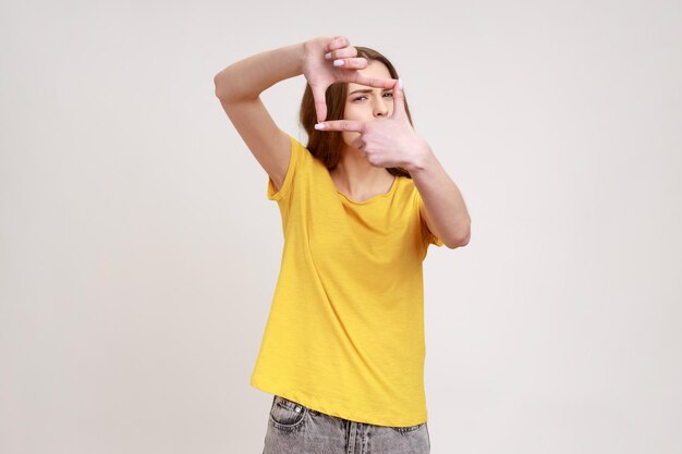 Winsome vrouw van jonge leeftijd dragen gele casual Tshirt frame gebaar maken met haar vingers als ze door de camera compositie van foto kijkt Indoor studio shot geïsoleerd op grijze achtergrond