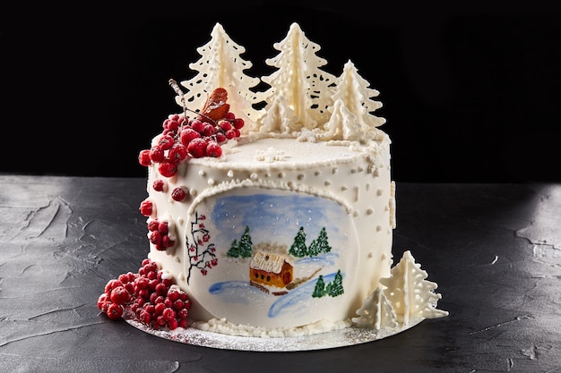 Photo winter cake covered with white icing with painted house in the forest trees and red rowan