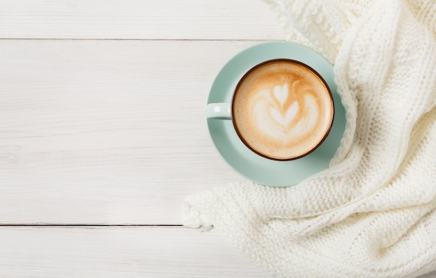 Winter cappuccino samenstelling. Blauwe koffiekopje bovenaanzicht met schuim en gebreide trui aan witte houten tafel. Warme dranken concept opwarmen