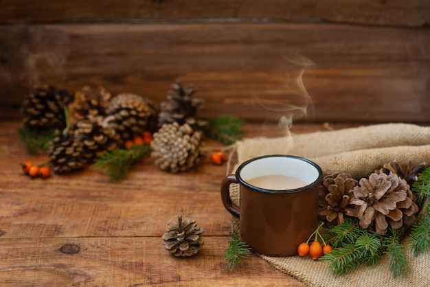 素朴なスタイルの冬、クリスマスの背景。松ぼっくり、トウヒの枝、ローズヒップの間の木製の表面にあるテーブルクロスの上に、ホットミルクティーが入った金属製のヴィンテージマグカップが立っています。コピースペース、フラットレイ