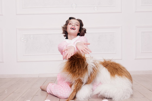 Winter, christmas, holiday, sweets, birthday, celebration and children concept- Cute little girl pajama with sweets sitting on the floor, happy childhood concept. girl gifts, soft dog, soap bubbles.