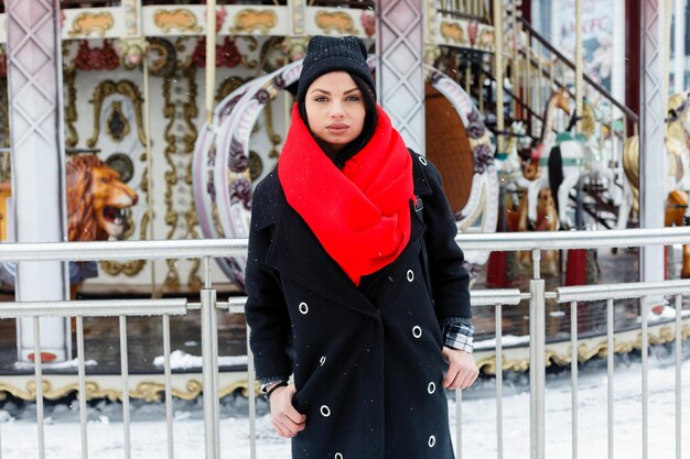 Winter, christmas, lifestyle, holidays and fashion concept - Pretty young smiling woman in black coat and red scarf posing in winter city. attractive brunette background of french merry-go-round